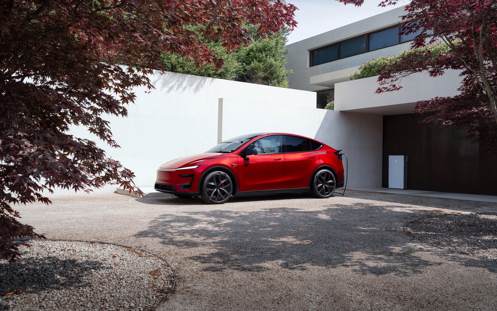 A red Tesla charging from a Tesla Wall Connector at the garage of a modern white house